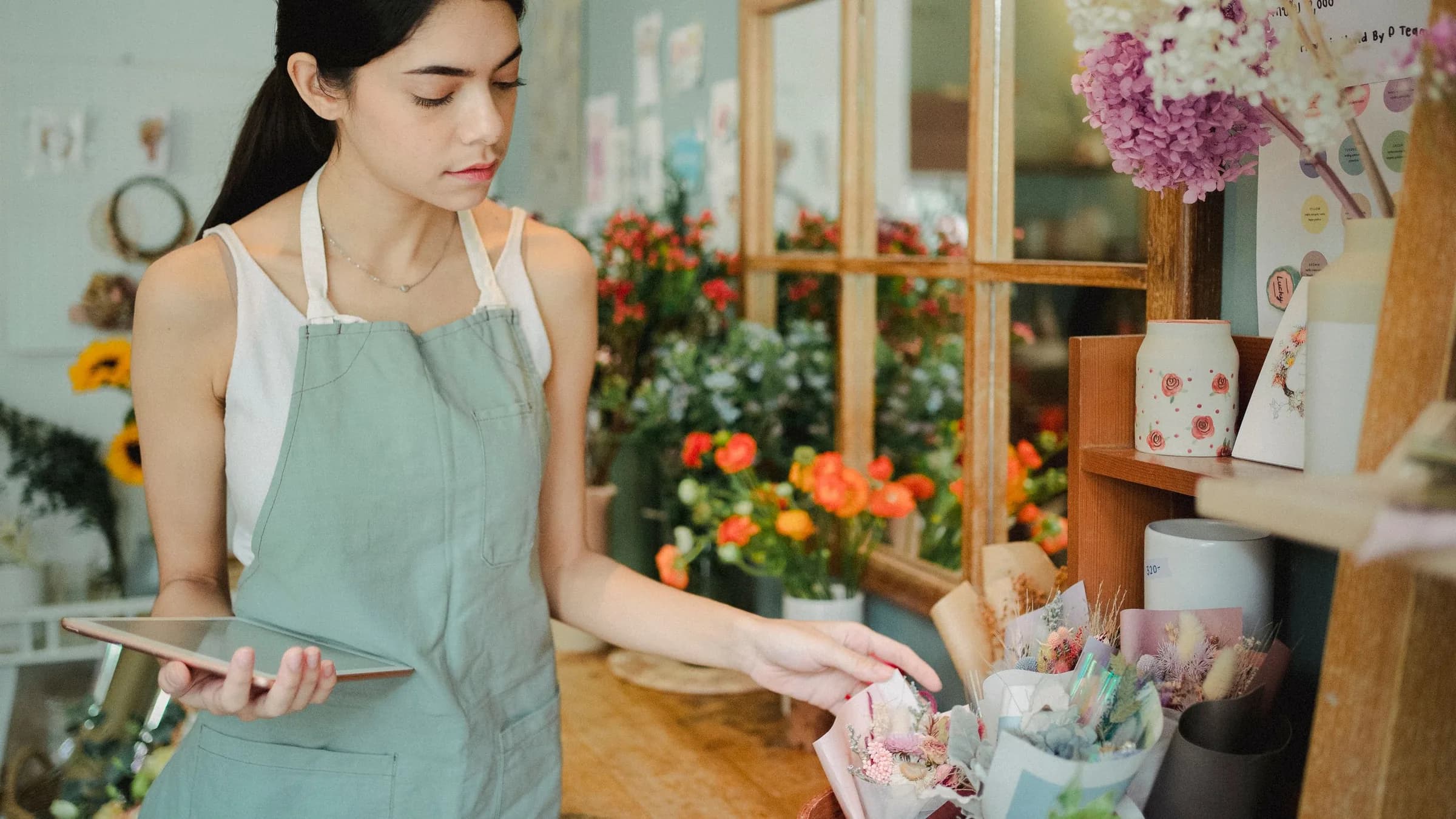 A florist in an apron holds a tablet while she checks a bouquet on the shop counter.