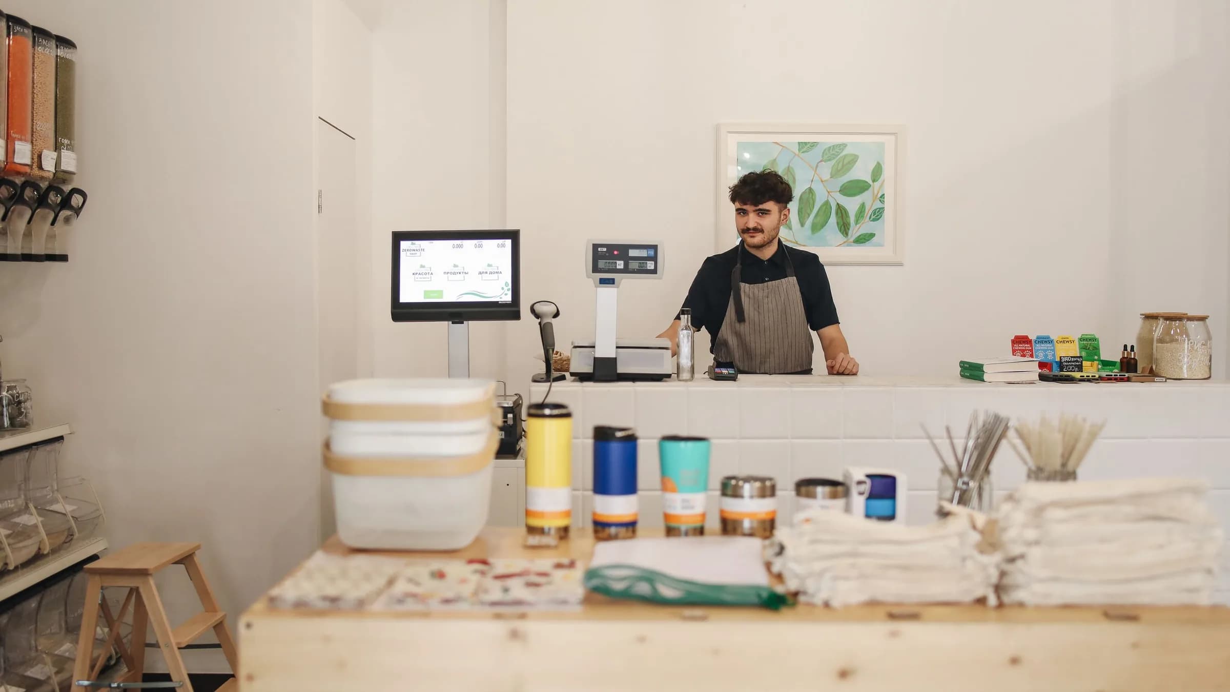 A shopkeeper stands behind a counter with a till screen, barcode scanner, scale and card reader.