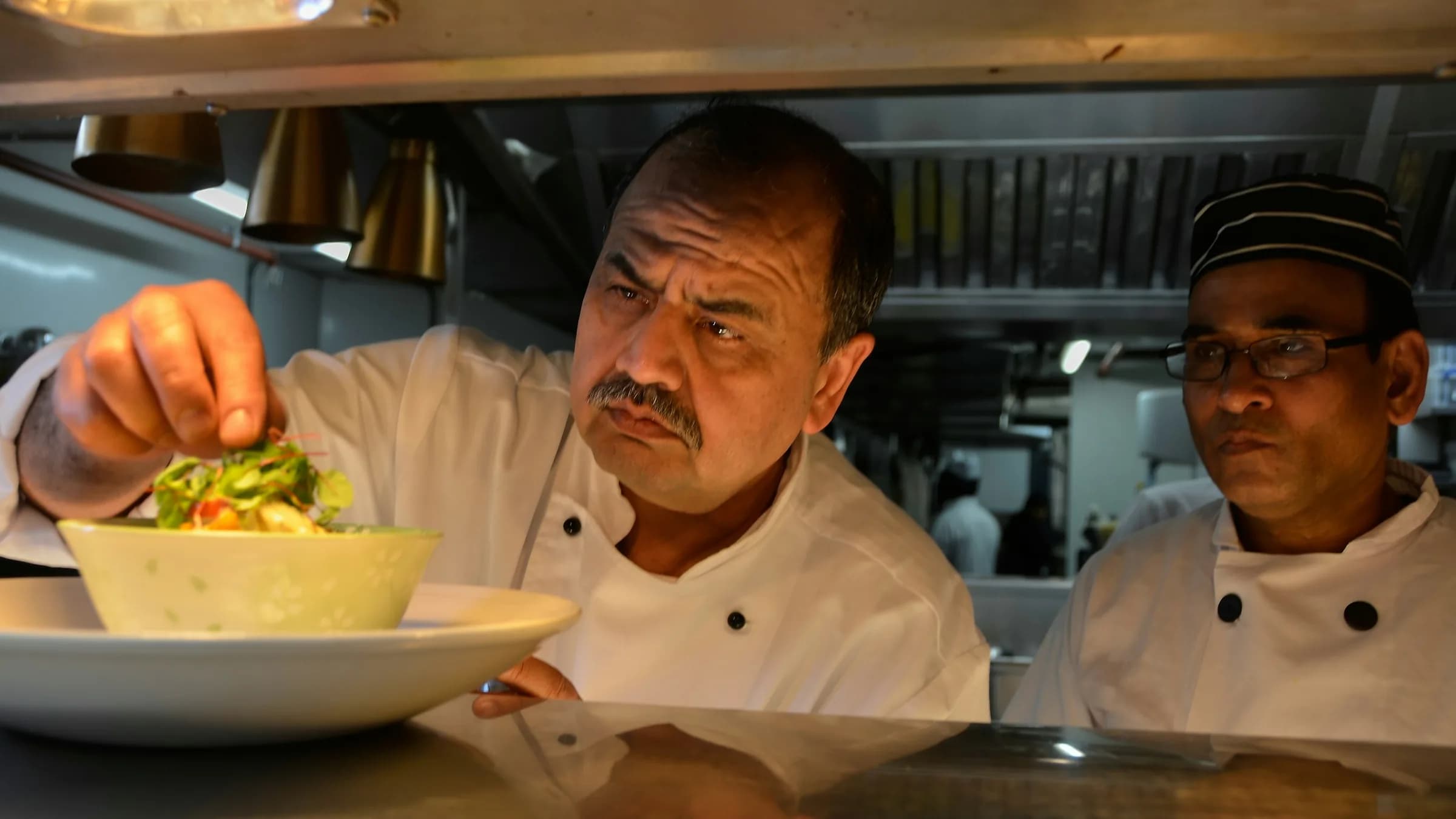 A chef garnishes a bowl at the kitchen pass while a second chef watches, steel shelves and extraction hoods behind them.
