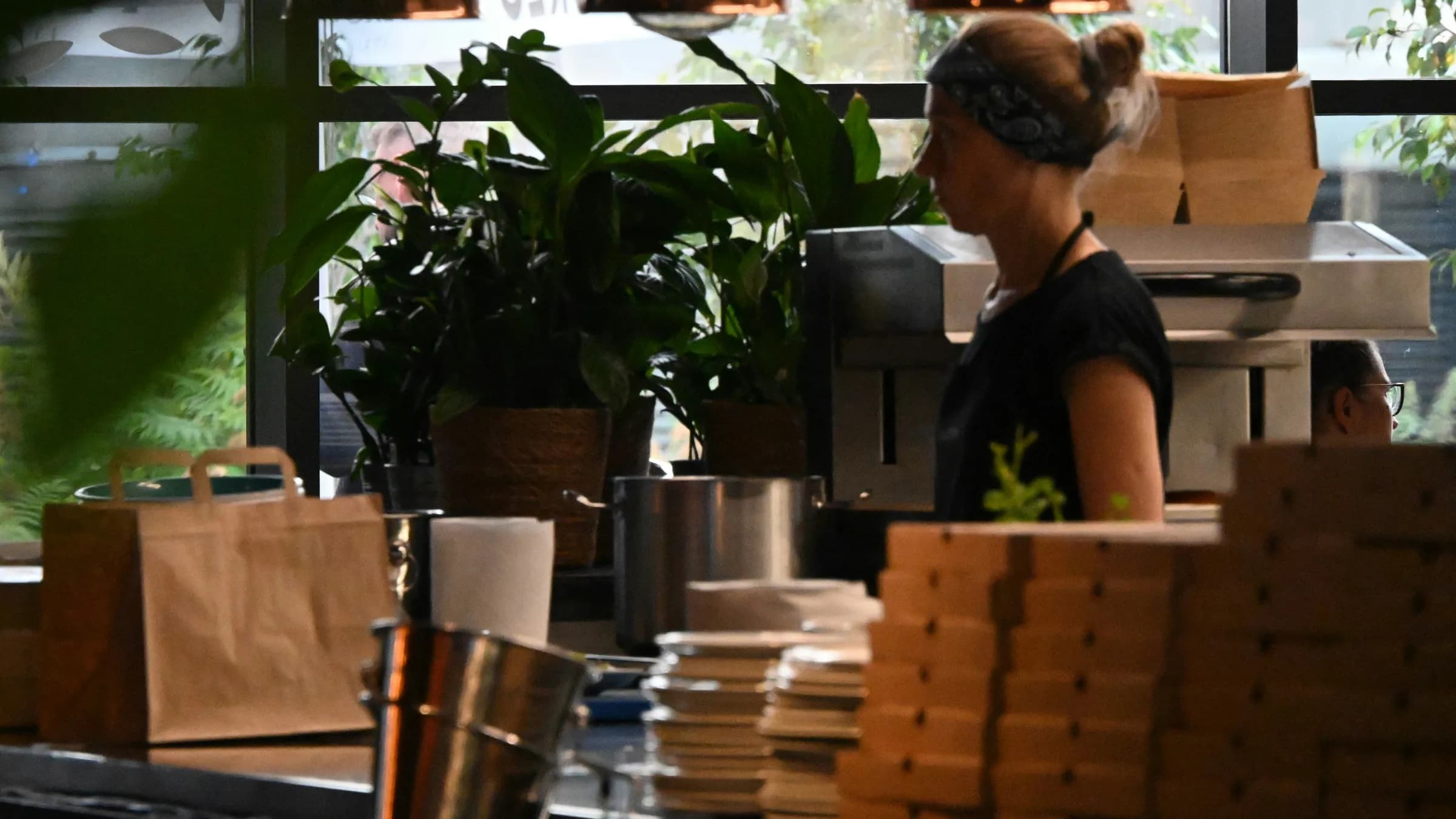 A cook works behind a takeaway counter stacked with brown paper bags and boxes ready for collection.