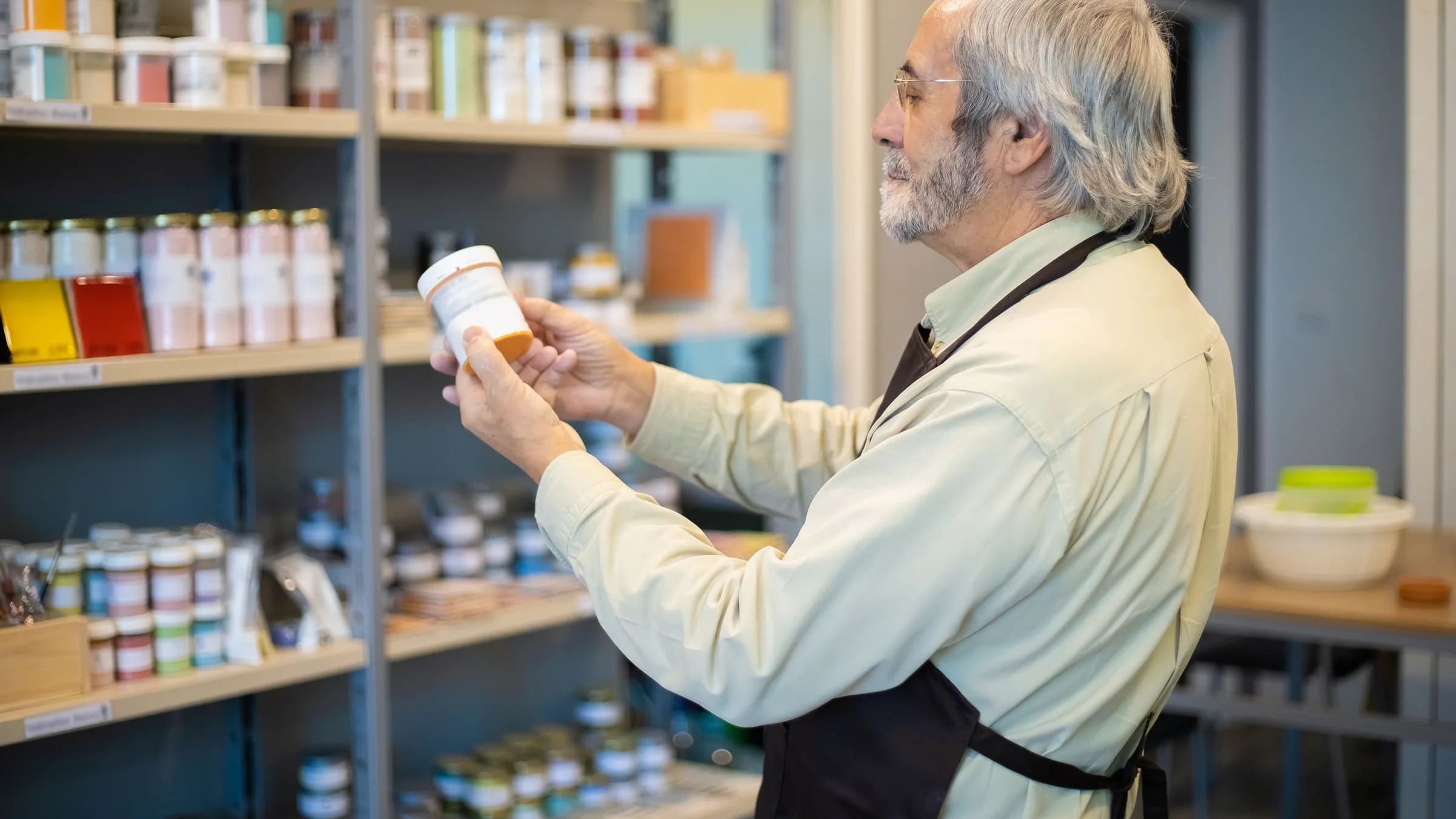 An older shopkeeper in an apron checks the label on a tub of paint in front of shelves of paint pots.