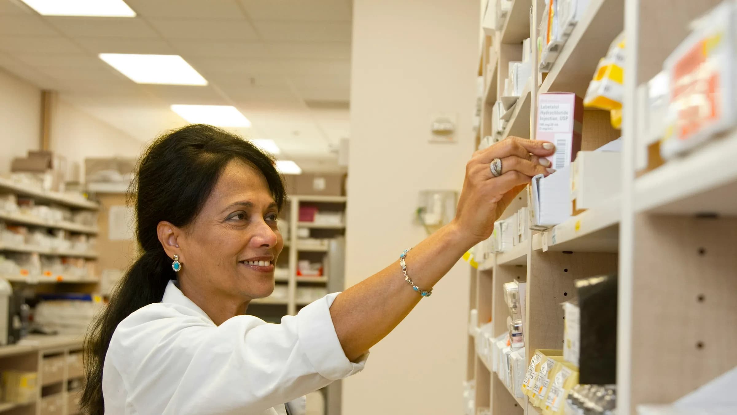 A smiling pharmacist reaches for a box on a dispensary shelf lined with medicine packs.