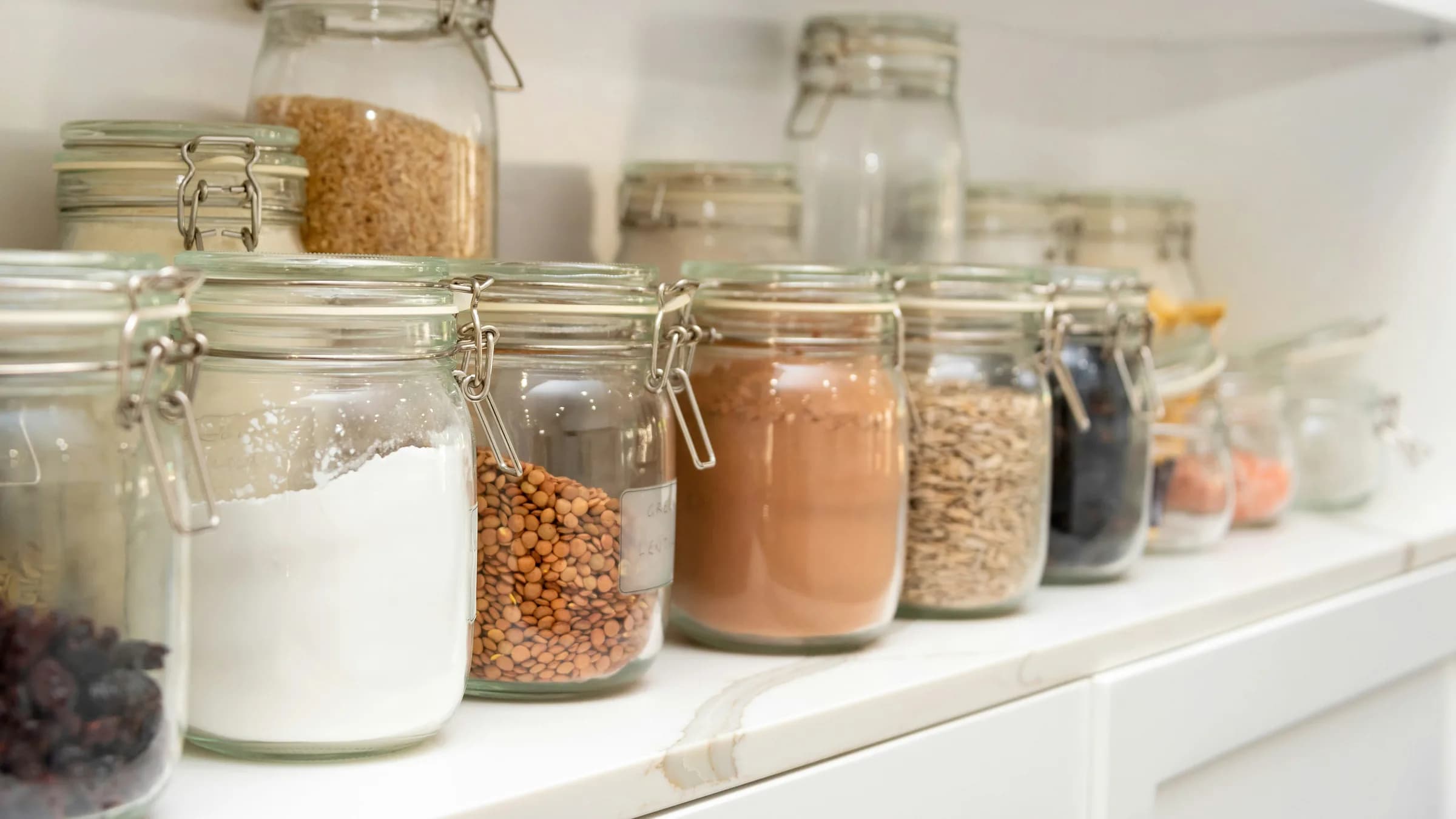 Glass storage jars of flour, lentils, cocoa and grains on a white shelf, some running low.