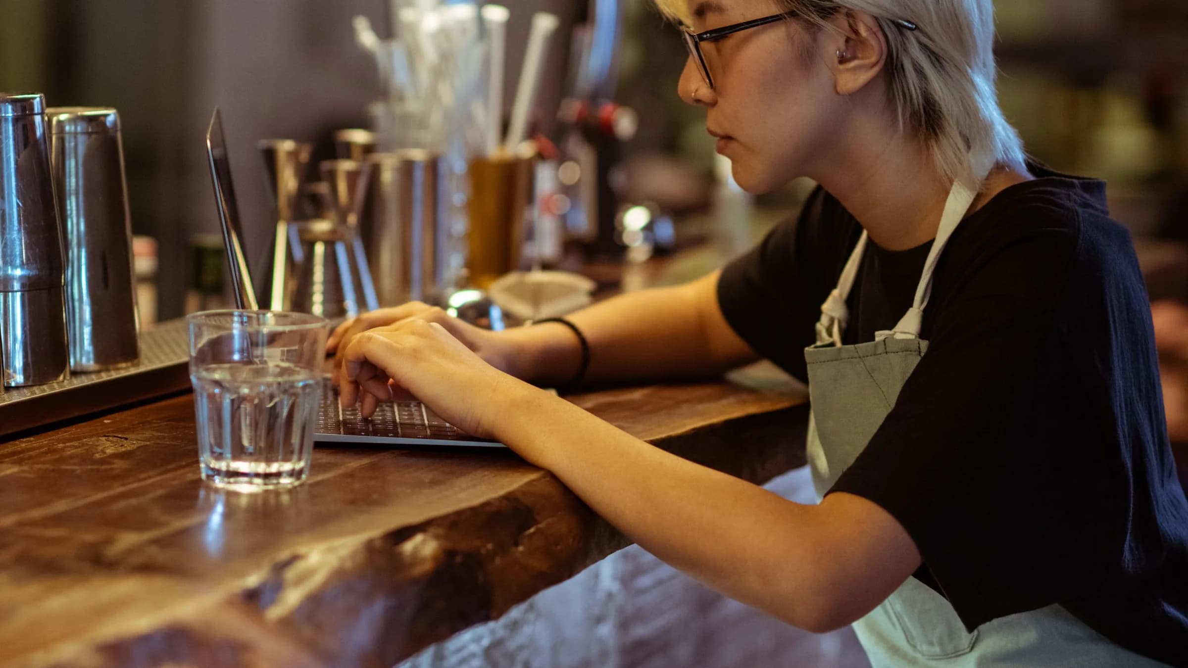 A bar worker in an apron types on a laptop at the end of a wooden counter, cocktail tools beside her.