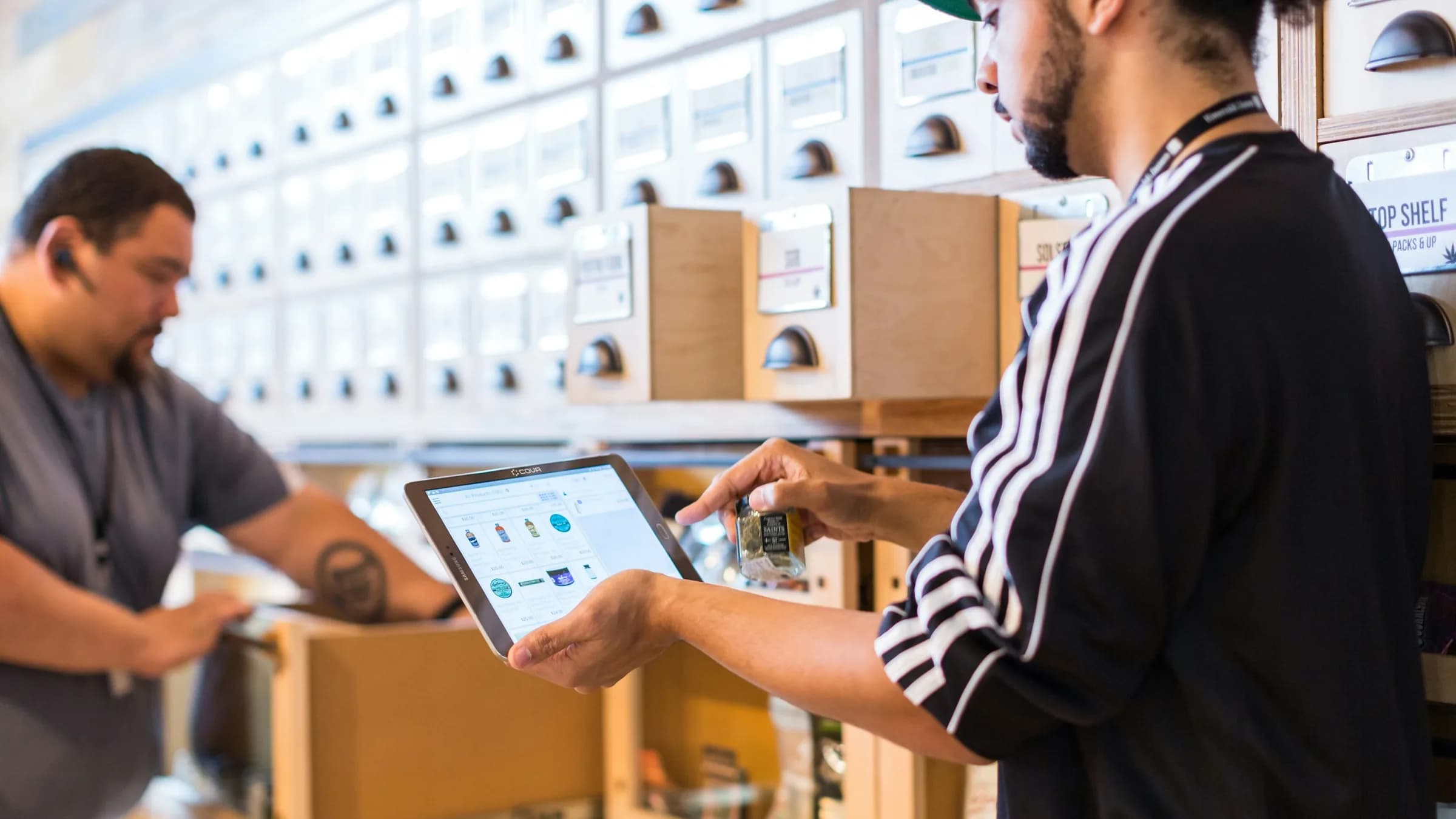 Two staff check stock in a storeroom, one holding a tablet showing product photos while the other opens a carton beside shelves of labelled boxes.