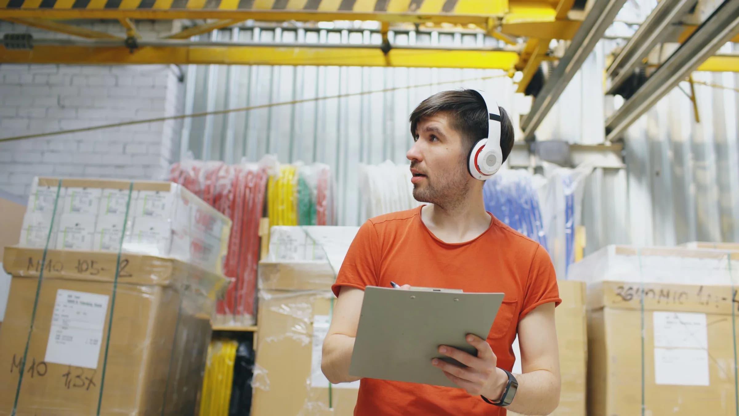 A warehouse worker with a clipboard checks stacked cartons and wrapped goods in a storeroom.