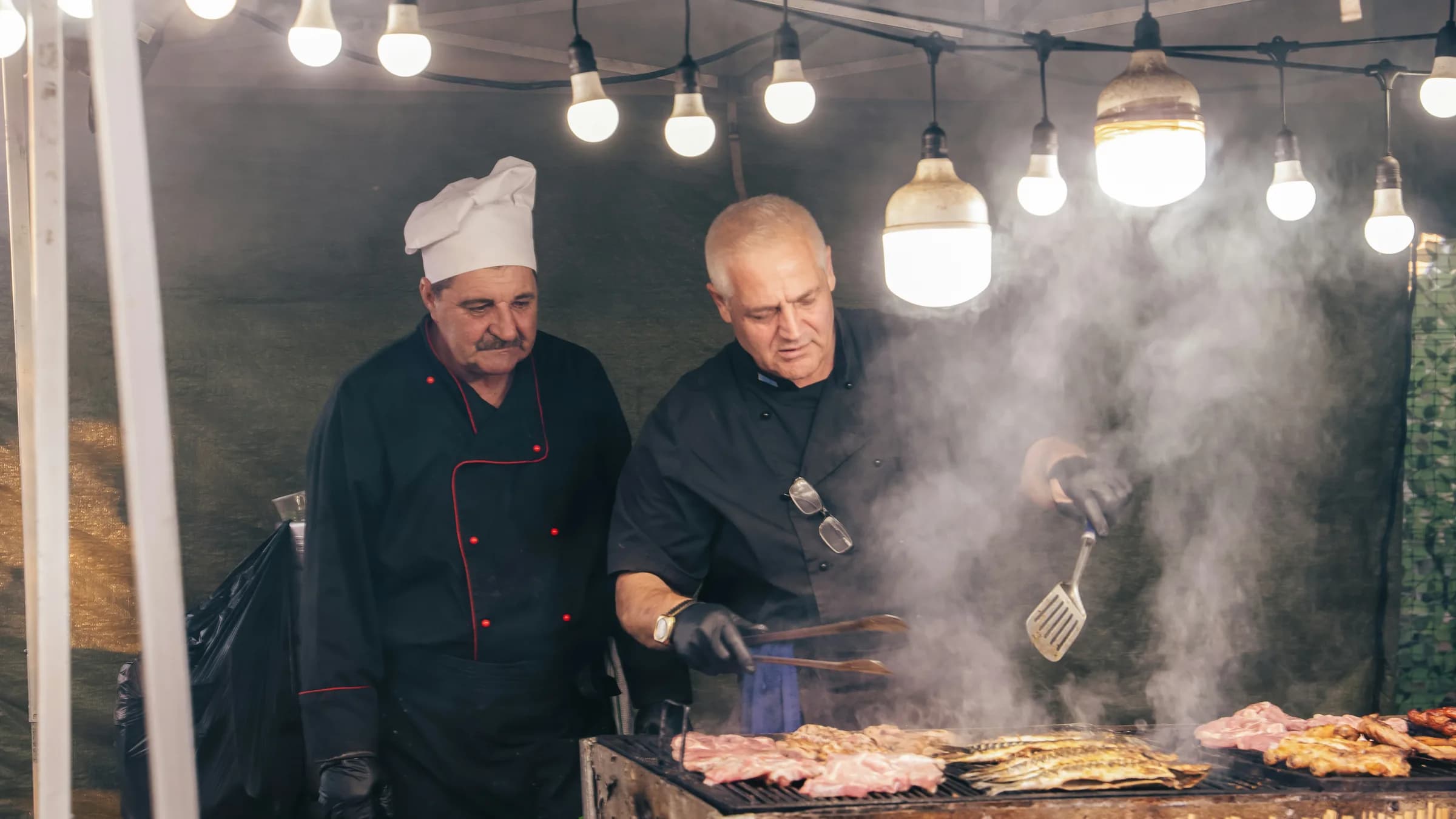 Two cooks grill meat and fish under a string of bulbs at an outdoor food stall.