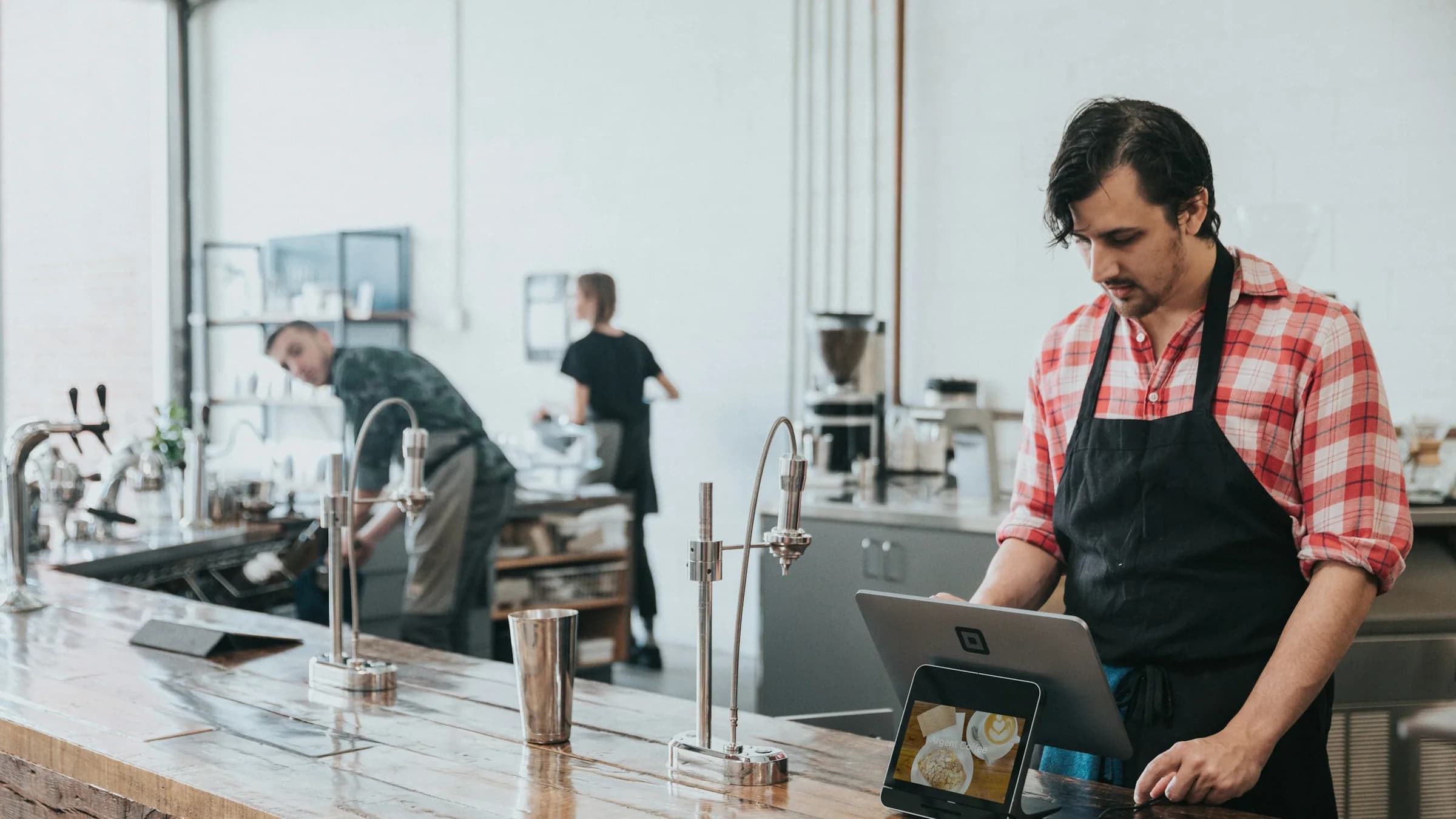A cafe worker in an apron uses a tablet till on a stand at the counter, with colleagues working behind him.