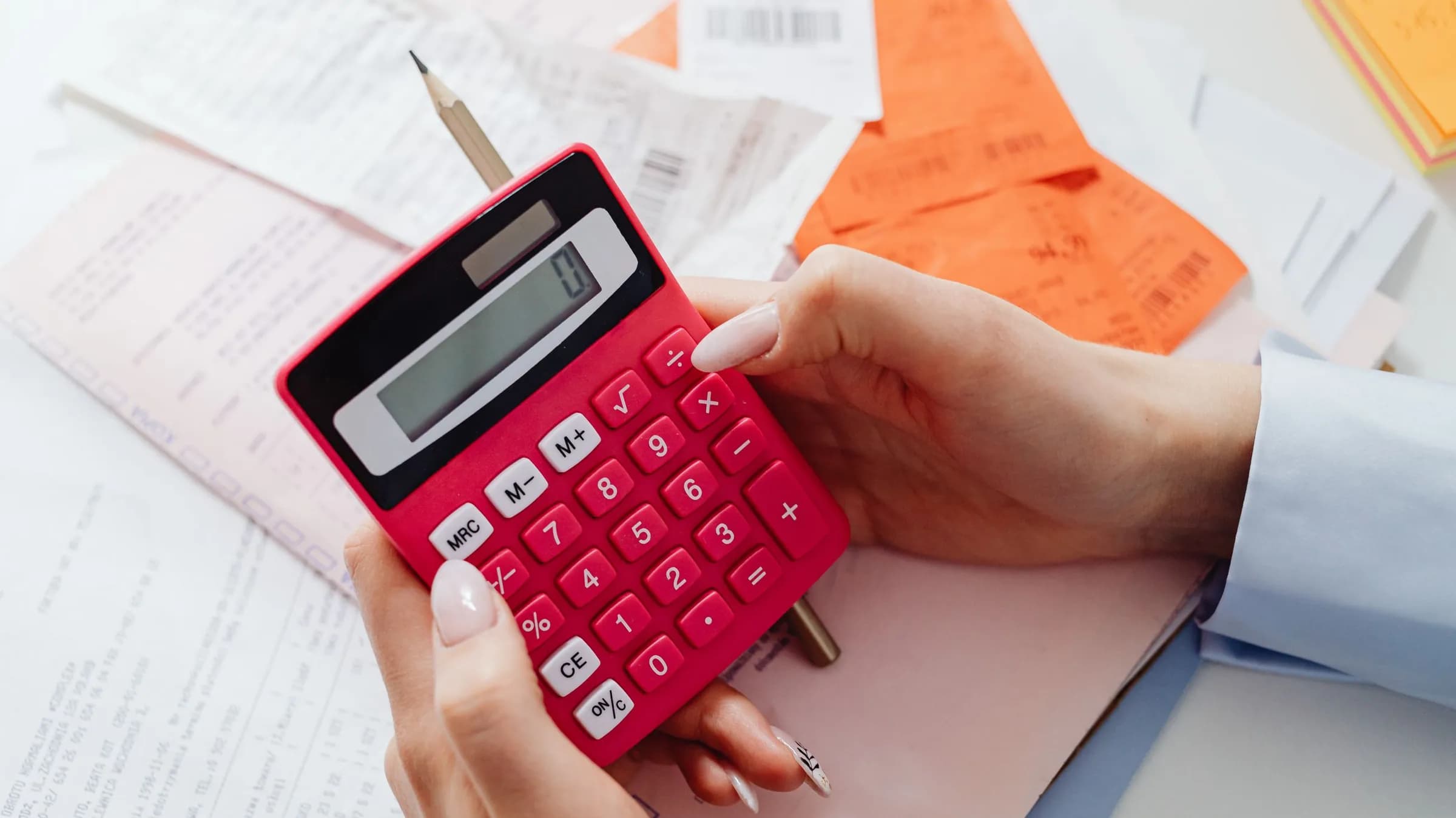 A hand holds a red calculator over printed invoices and orange receipt slips.